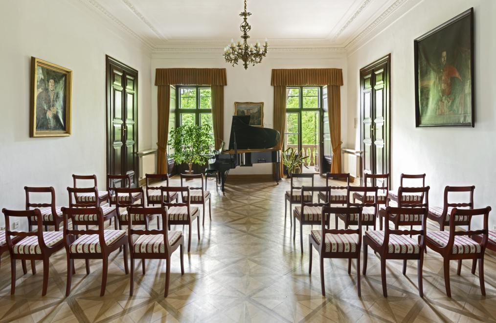 Fotografia wnętrza sali z rzędem krzeseł i fortepianem na tle dwóch dużych okien..