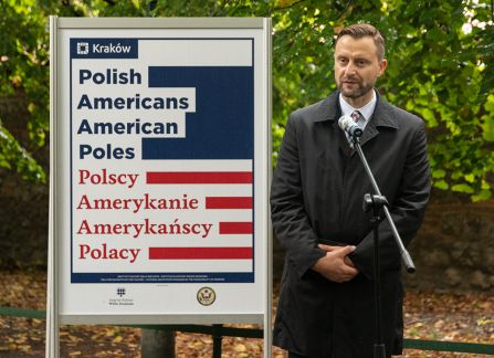 A photograph. An opening of the outdoor exhibition titled Polish Americans, American poles at planty in Krakow. President's plenipotentiary Robert piaskowski gives a speech standing next to the poster promoting the exhibition.
