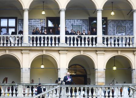 Fotografia. Fasada willi Decjusza. Mężczyzna w garniturze przemawia ze szczytu schodów. Na loggii pierwszego piętra ubrani na czarno członkowie chóru.
