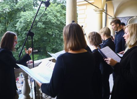 Fotografia. Plenerowy występ muzyczny chóru. Ubrani na czarno chórzyści śpiewają stając u szczytu schodów przed willą Decjusza