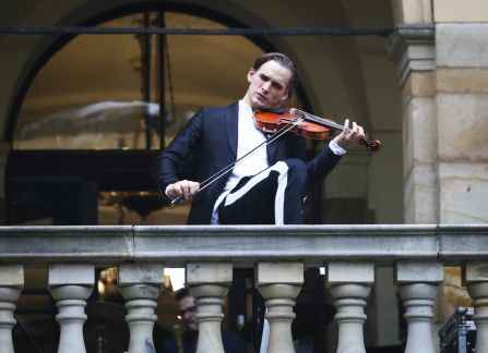 Fotografia. Plenerowy występ muzyczny. mężczyzna w garniturze gra na skrzypcach. W tle łuki arkad fasady Willi Decjusza.