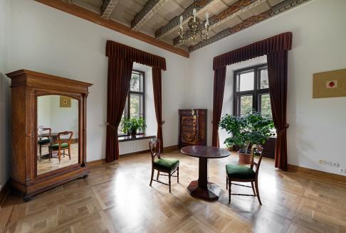 fotografia: wnętrze sali Willi Decjusza. Na zdjeciu widać zabytkowy stół i dwa krzesła oraz drewnianą szafę z lustrem i dwa okna oraz fragment belkowanego sufitu.