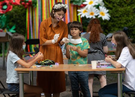 Fotografia. Kobieta w brązowej sukience prowadzi warsztaty plastyczne dla dzieci. W tle ogród, kolorowe dekoracje.