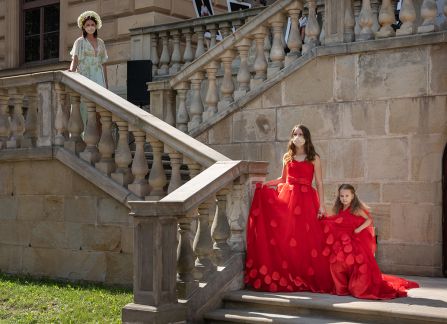 Fotografia. Trzy młode kobiety w długich, czerwonych i białych sukniach stoją na schodach przed Willą Decjusza.