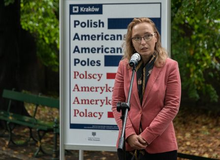 A photograph. An opening of the outdoor exhibition titled Polish Americans, American poles at planty in Krakow. the director of the Villa Decius institute for culture Dominika Kasprowicz gives a speech standing next to the poster promoting the exhibition.