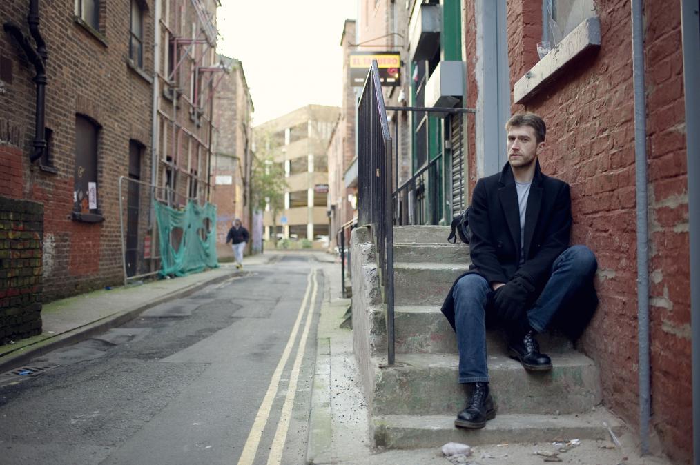 portrait picture of UNESCO resident Mark Pajak. A man is sitting on the low stair, a narrow city street in the background.