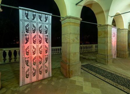 a photograph. large-format cutout with a traditional pattern, displayed between the columns of arcades on the facade of the Decius villa. Photo taken after dark, the cut-out highlighted in green and red.